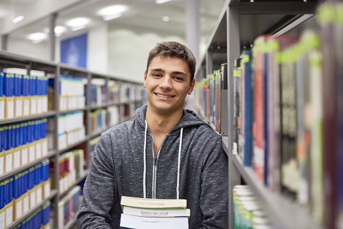 A male student wearing a grey hoodie, stood in front of the library shelves holding books. 