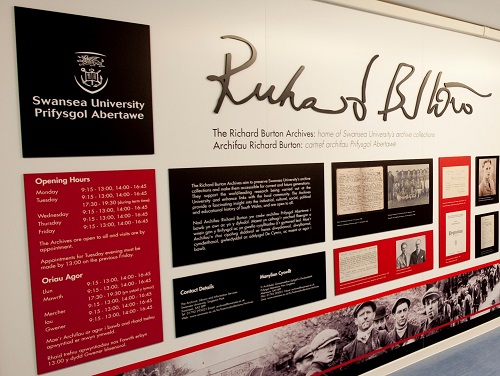 View of the white, red, and black wall outside Richard Burton Archives entrance hallway which contains images from the collection and information such as opening hours.